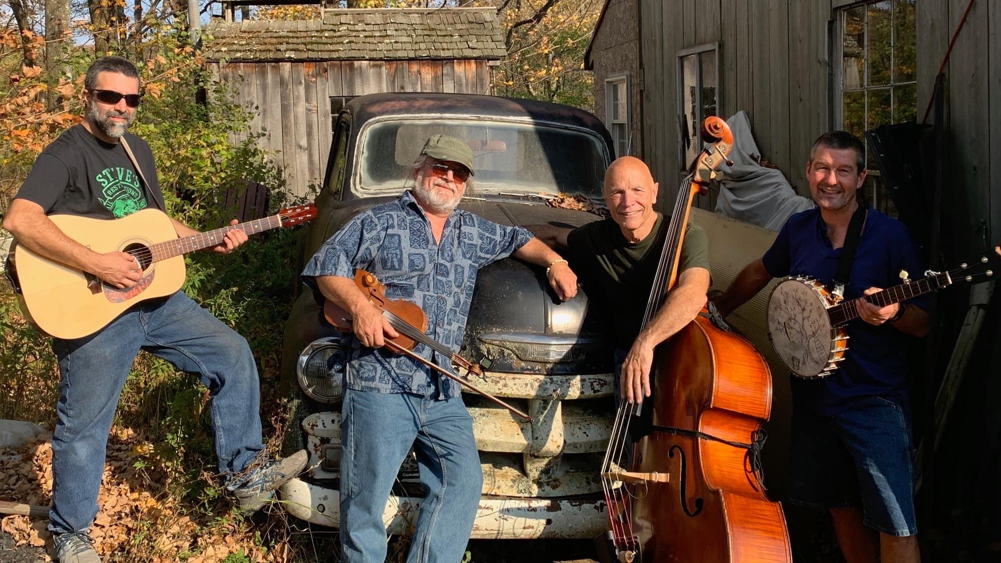 Four older white men stand in front of an old truck and rustic shack. They are holding instruments including a guitar, fiddle, stand-up bass, and banjo
