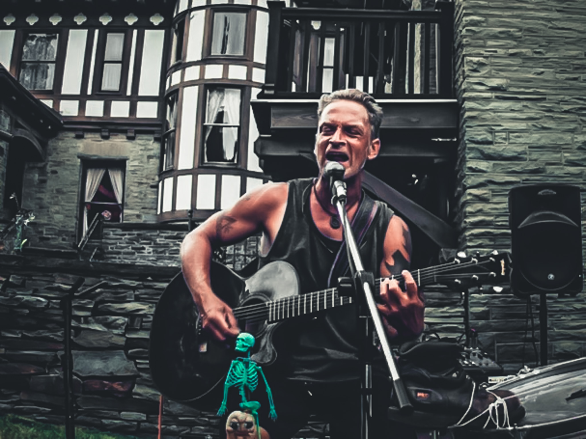 A white man sings into a mic in front of a stone building. He is playing a guitar and a small, bright green skeleton is in front of him