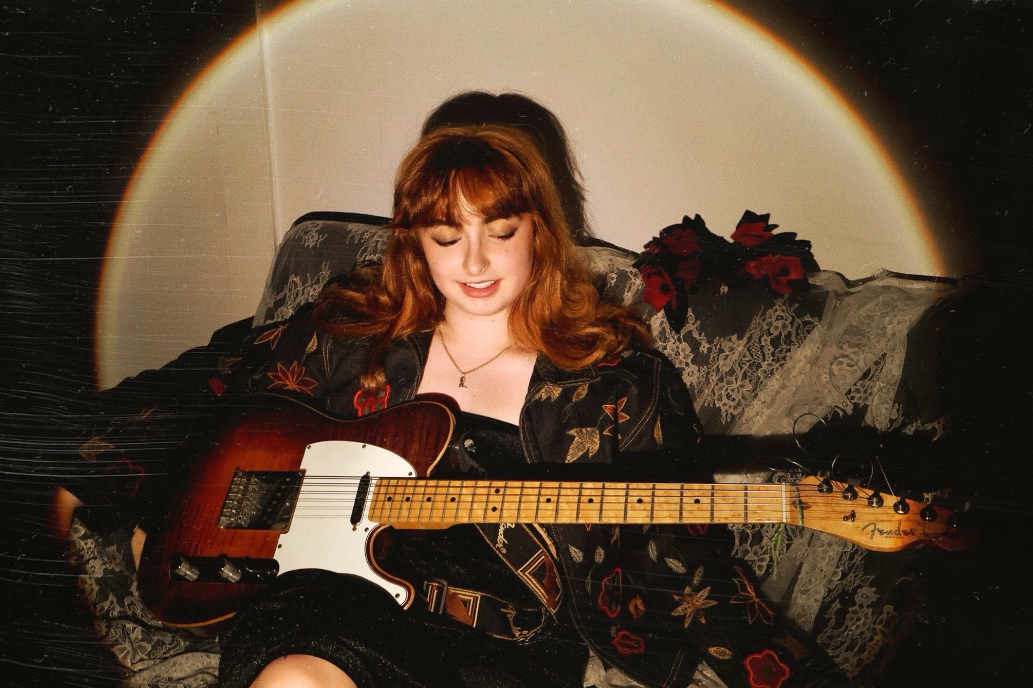 A young white woman with red hair seated on a lace covered couch and holding an electric Fender guitar