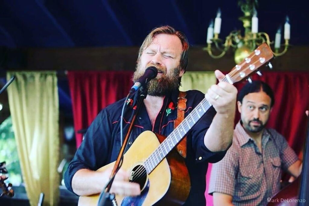 A white man with a beard plays an acoustic guitar. His eyes are closed as he sings into a microphone.